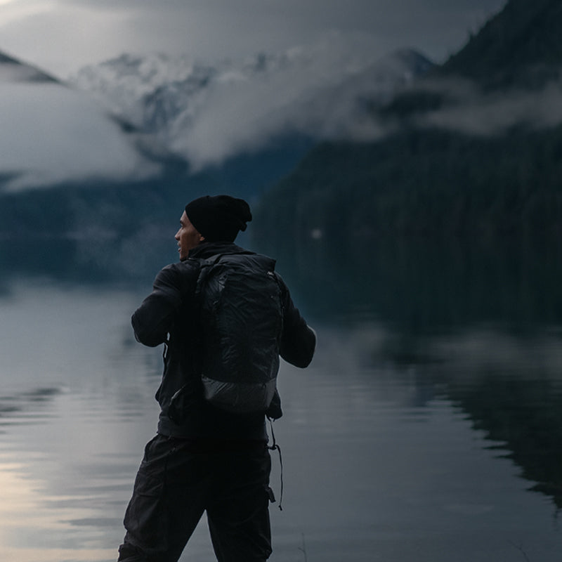 A person in a black beanie and backpack stands by a calm lake, surrounded by misty mountains and a cloudy sky.